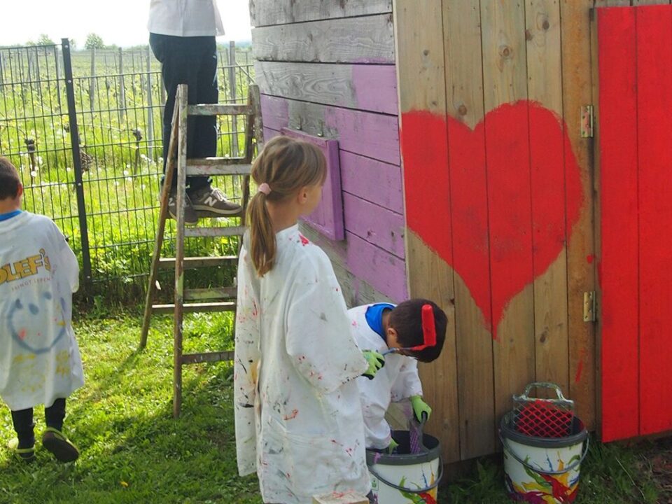 Blondes Mädchen in Malerkittel steht vor einer Holzhütte und malt ein großes rotes Herz darauf